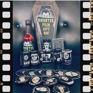A themed horror movie gift set from Film Photography Project arranged on a dark surface against a black background, featuring a tall coffin-shaped sign reading 'Monster Film Gift Box,' a bottle of monster hot sauce and matching label, 6 rolls of film, stickers and buttons. The designs are black and white with illustrated monster faces and text such as 'The Wolf,' 'Dracula,' 'Frankenstein,' and 'Hydro Hilarity.